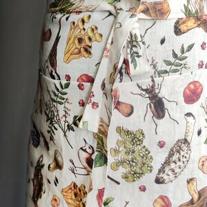 May include: Off-white apron with a botanical print featuring mushrooms, insects, berries, and foliage. The apron has a wrap-around design with a tie closure and two front pockets. The print includes shades of brown, yellow, green, and red.