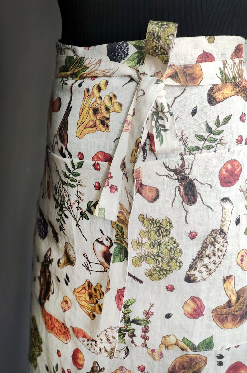 May include: Off-white apron with a botanical print featuring mushrooms, insects, berries, and foliage. The apron has a wrap-around design with a tie closure and two front pockets. The print includes shades of brown, yellow, green, and red.