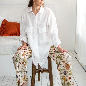 May include: A white linen shirt with a relaxed fit and a button-up front. The shirt has two large pockets on the front. The model is wearing the shirt with a pair of white trousers with a colourful floral print featuring mushrooms, berries, and leaves.