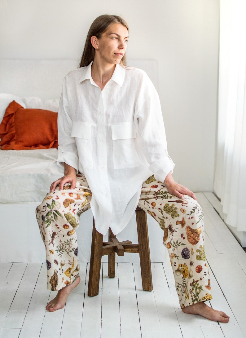 May include: A white linen shirt with a relaxed fit and a button-up front. The shirt has two large pockets on the front. The model is wearing the shirt with a pair of white trousers with a colourful floral print featuring mushrooms, berries, and leaves.