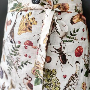 May include: Off-white apron with a botanical print featuring mushrooms, insects, and foliage. The apron has a wrap-around design with a matching tie. The print includes various colors, such as brown, green, and red, creating a detailed and naturalistic design.