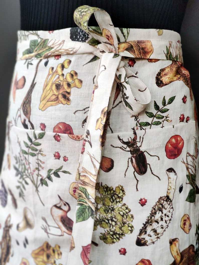May include: Off-white apron with a botanical print featuring mushrooms, insects, and foliage. The apron has a wrap-around design with a matching tie. The print includes various colors, such as brown, green, and red, creating a detailed and naturalistic design.