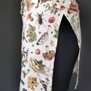 May include: A wrap-around apron with a beige background and a botanical print featuring mushrooms, insects, and plants. The apron has a tie closure and a split design, offering coverage and style. The print includes various colors.