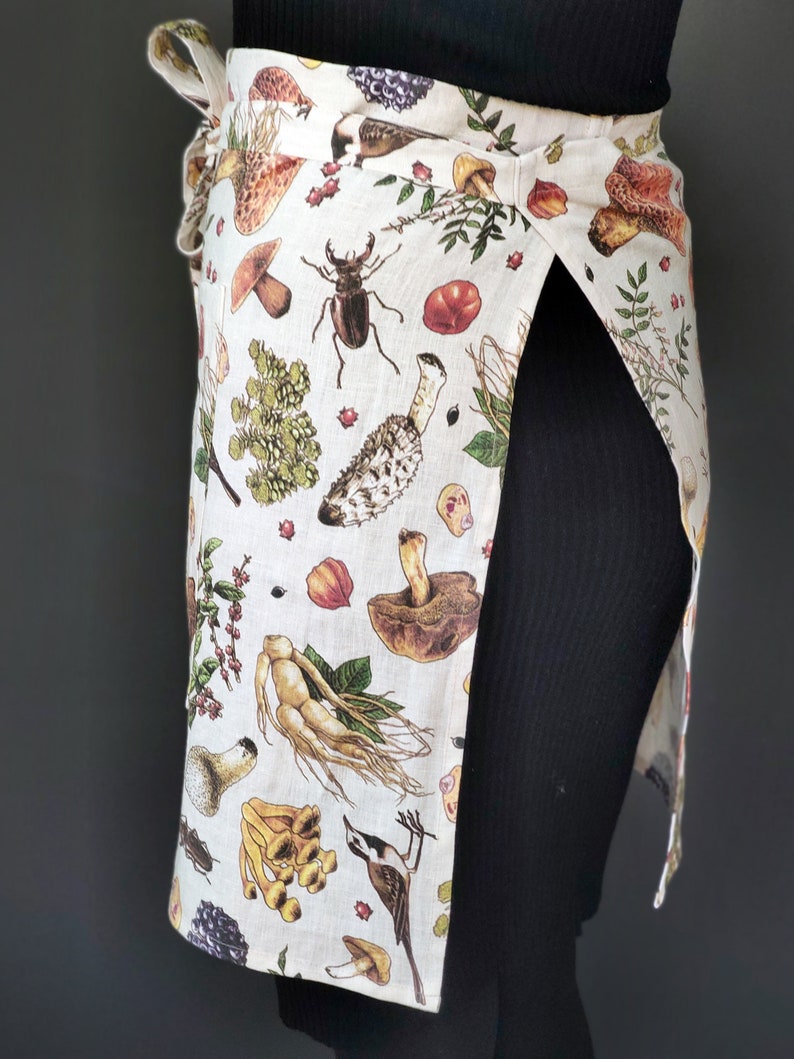 May include: A wrap-around apron with a beige background and a botanical print featuring mushrooms, insects, and plants. The apron has a tie closure and a split design, offering coverage and style. The print includes various colors.