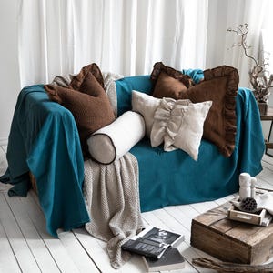 Könnte beinhalten: Ein türkisfarbenes Sofa ist mit verschiedenen Kissen und Decken geschmückt. Braune, beige und weiße Kissen in verschiedenen Formen und Texturen sind auf dem Sofa angeordnet. Eine beige Decke und ein weißes Nackenkissen ergänzen das Ensemble. Das Sofa steht auf einem weißen Holzboden.