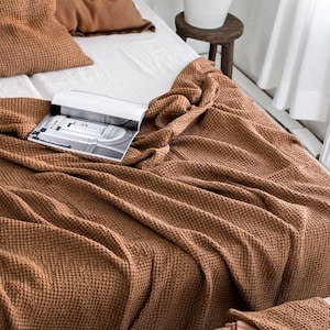 May include: A brown waffle weave throw blanket is draped over a white bed with brown pillows. The blanket is folded and arranged in a decorative way. A book is lying on top of the blanket.
