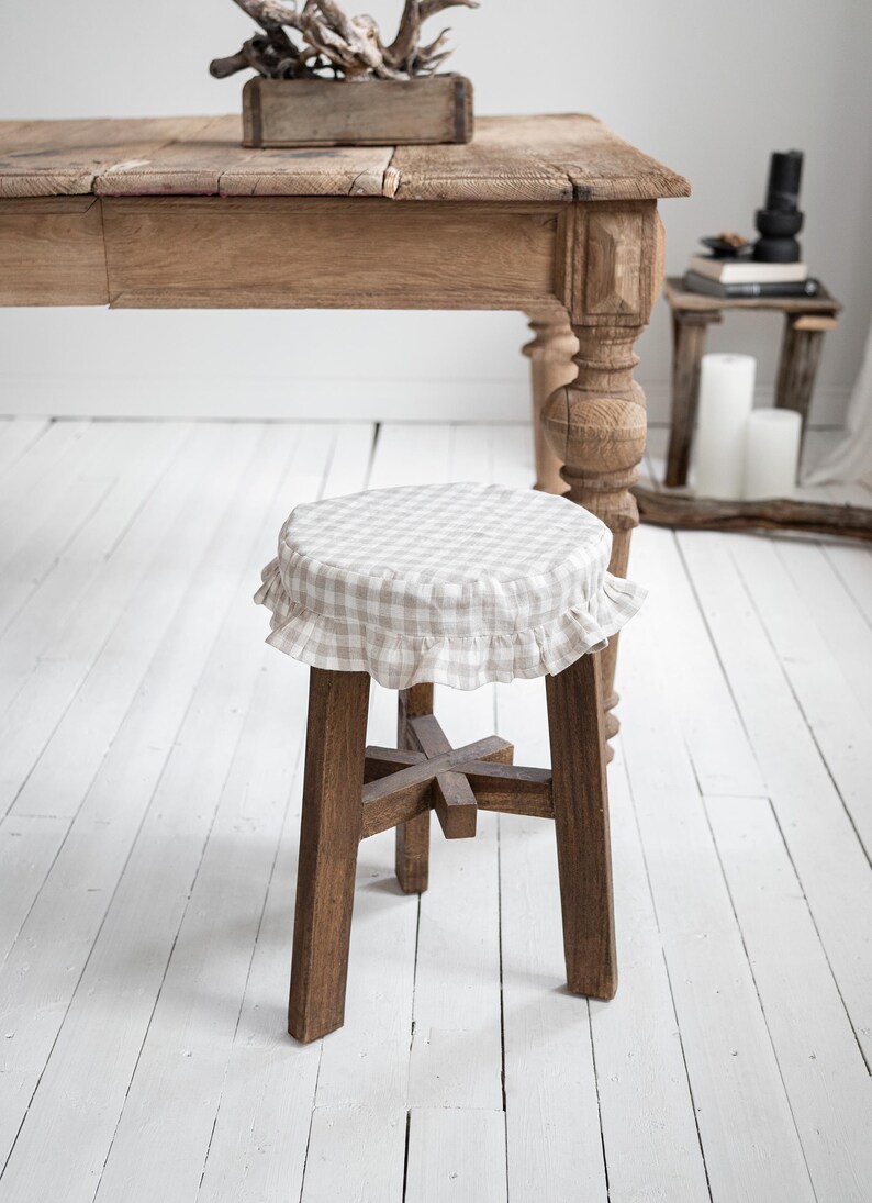 Pu&ograve; includere: Sgabello in legno con rivestimento a quadretti e finiture a balze. Lo sgabello ha una base a quattro gambe ed &egrave; realizzato in legno scuro. Il rivestimento della seduta presenta un motivo a quadretti beige chiaro e bianco. Lo sgabello &egrave; in una stanza con un tavolo di legno.