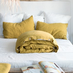 Peut inclure: Une housse de couette et des taies d'oreiller en lin jaune moutarde sur un lit blanc avec une tête de lit en bois. La housse de couette est pliée et comporte deux boutons sur le côté. Il y a un magazine sur un banc en bois au premier plan.