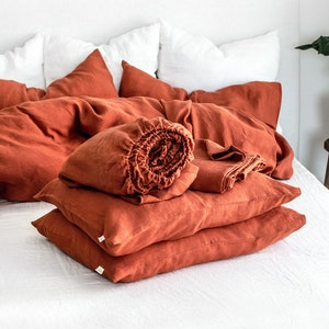 May include: A stack of rust-coloured linen pillowcases and a rolled-up linen duvet cover on a white bed.