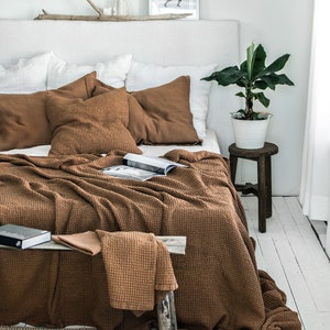 May include: A brown waffle weave throw blanket draped over a bed with white linen bedding and brown throw pillows. The blanket is folded over a wooden stool with a potted plant on top.