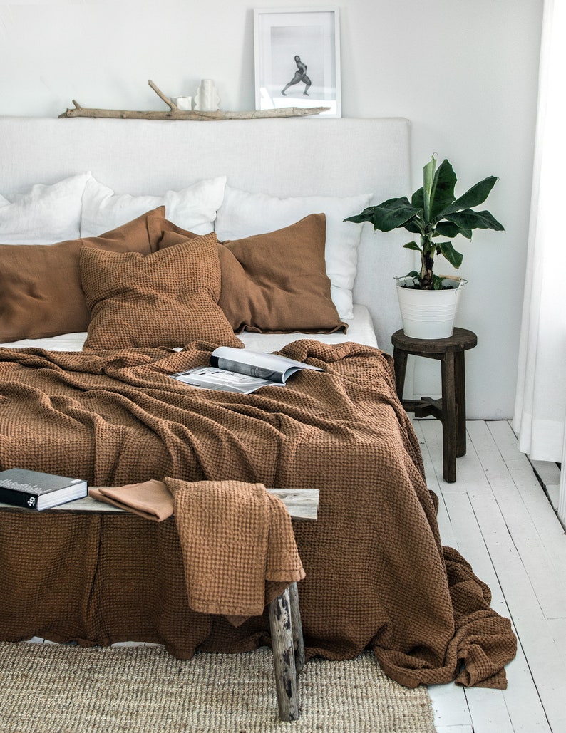May include: A brown waffle weave throw blanket draped over a bed with white linen bedding and brown throw pillows. The blanket is folded over a wooden stool with a potted plant on top.