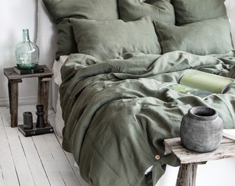 Op de afbeelding: Een groene linnen dekbedovertrek met bijpassende kussenslopen op een wit bed. Het dekbedovertrek heeft knopen en banden aan de onderkant. Het bed staat in een slaapkamer met witte muren en houten vloeren.