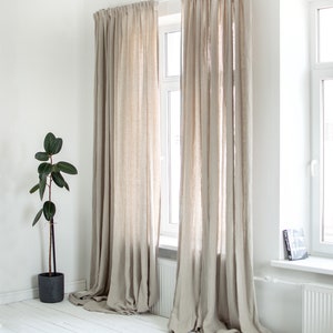May include: Two pairs of beige linen curtains hanging in a window. The curtains are gathered at the top and hang to the floor. The windows are white and have white trim.