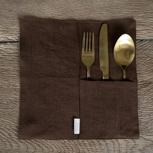 May include: A brown linen cutlery holder with a pocket containing a gold fork, knife, and spoon. The cutlery has black handles. The holder has a small white tag that reads "GRANNET HOME".