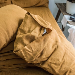 Puede incluir: Primer plano de un juego de cama de lino amarillo mostaza con una almohada doblada encima. La ropa de cama est&aacute; arrugada y tiene un aspecto natural y texturizado.