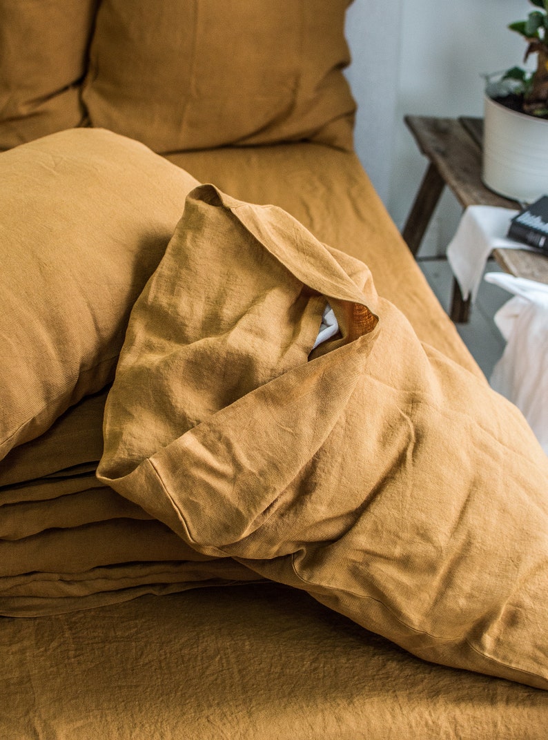Puede incluir: Primer plano de un juego de cama de lino amarillo mostaza con una almohada doblada encima. La ropa de cama est&aacute; arrugada y tiene un aspecto natural y texturizado.