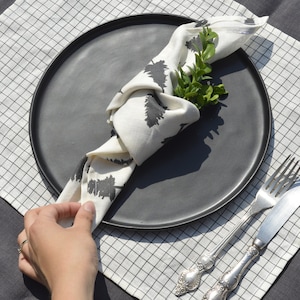 Puede incluir: Un plato negro con una servilleta blanca doblada en un nudo decorativo. La servilleta tiene un patrón de árboles en blanco y negro. Una ramita de vegetación se coloca encima de la servilleta. El plato está sobre un mantel individual a cuadros blanco y negro.