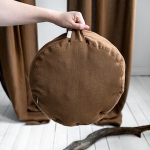 May include: A round, brown cushion with a zipper closure and a small fabric tag. The cushion is held up by a hand, with brown curtains in the background. A piece of wood is on the white floor.