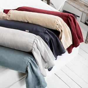 May include: A collection of linen bolster pillows in various colors, including red, beige, navy, light gray, and blue. Each pillow has a ruffled edge. The pillows are arranged on a white bed, with a wooden bench and other decor in the background.