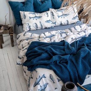 May include: A bed with a white headboard, adorned with blue pillows and bedding. The duvet cover and pillowcases feature a fish pattern in shades of blue on a white background. A dark blue textured blanket is draped over the bed.