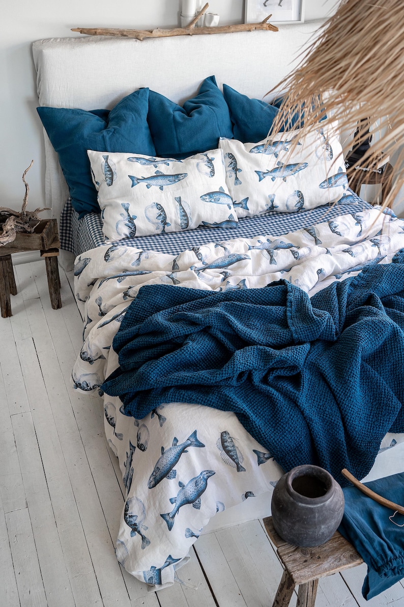 May include: A bed with a white headboard, adorned with blue pillows and bedding. The duvet cover and pillowcases feature a fish pattern in shades of blue on a white background. A dark blue textured blanket is draped over the bed.