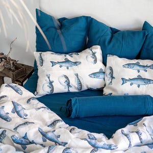 May include: A bed with teal blue bedding and pillows. The pillows and duvet feature a white background with blue fish illustrations. A blue bolster pillow and a blue knitted blanket are also visible. The bed is against a white headboard.