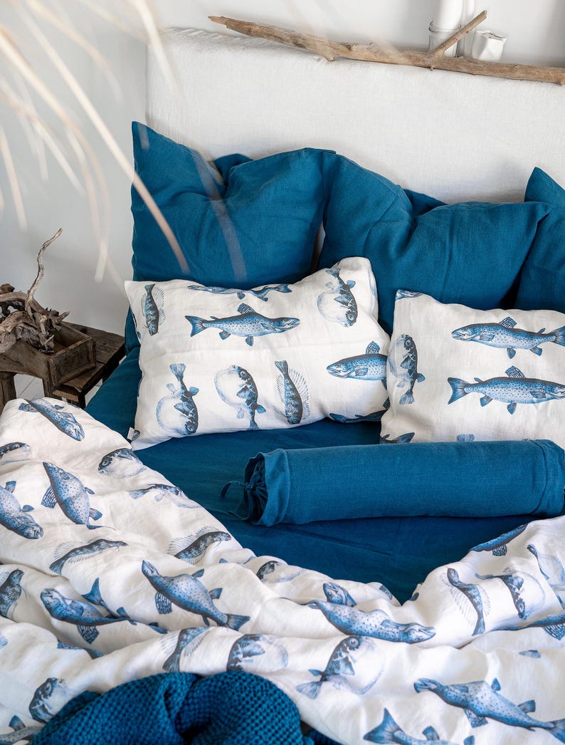 May include: A bed with teal blue bedding and pillows. The pillows and duvet feature a white background with blue fish illustrations. A blue bolster pillow and a blue knitted blanket are also visible. The bed is against a white headboard.
