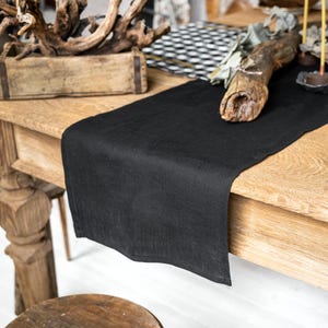 May include: A solid black rectangular table runner made of fabric, draped over the edge of a light brown wooden table. The runner has a smooth, matte texture and clean, straight edges, extending across the surface of the wooden furniture piece.