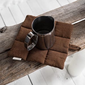 May include: A stainless steel pitcher sits on a brown quilted square hot pad. The hot pad is on a weathered wooden surface. A white bottle is in the background. The hot pad has a tag that reads "GRANNET HOME".