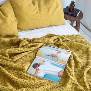 May include: A mustard yellow waffle weave throw blanket on a white bed with a magazine on top. The blanket is folded in half and the magazine is open to a page with a photo of a person wearing a white shirt.