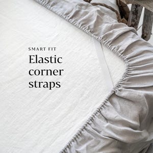 Pode incluir: Close-up de um lençol de elástico cinza com tiras elásticas nos cantos. O lençol tem um fundo branco com o texto "SMART FIT Elastic corner straps" impresso nele.