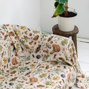May include: A beige linen throw blanket with a mushroom and forest-themed print. The blanket is draped over a couch and features illustrations of mushrooms, leaves, and insects.