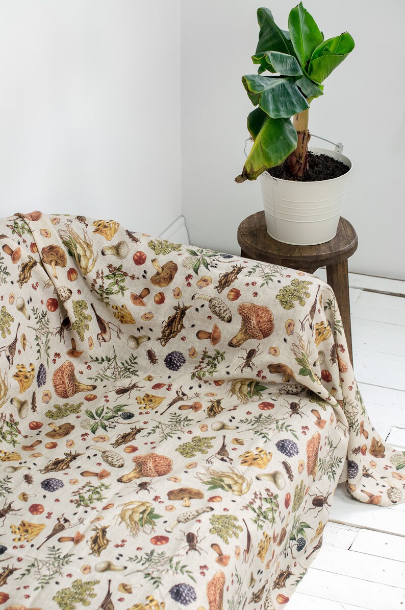 May include: A beige linen throw blanket with a mushroom and forest-themed print. The blanket is draped over a couch and features illustrations of mushrooms, leaves, and insects.