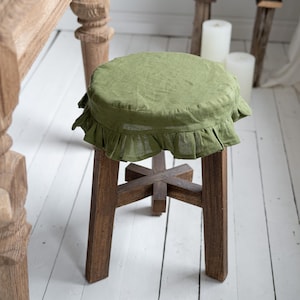 Pu&ograve; includere: Uno sgabello in legno con una fodera per sedile in lino verde oliva. La fodera ha un bordo a balze. Le gambe dello sgabello sono in legno marrone scuro e sono unite da un supporto a forma di croce.