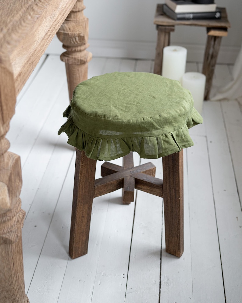 Pu&ograve; includere: Uno sgabello in legno con una fodera per sedile in lino verde oliva. La fodera ha un bordo a balze. Le gambe dello sgabello sono in legno marrone scuro e sono unite da un supporto a forma di croce.