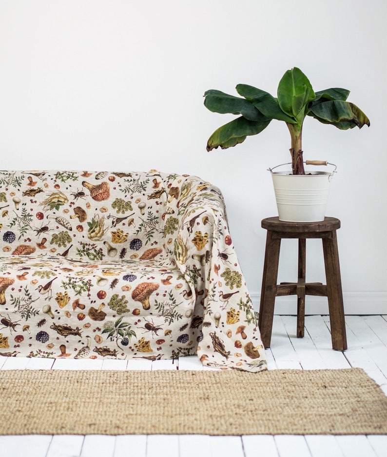 May include: A white couch with a throw blanket featuring a mushroom and forest-themed print. The blanket has a beige background with brown, green, and red accents. A potted plant with green leaves sits on a wooden stool next to the couch.