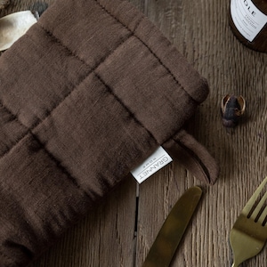 May include: A close-up of a brown quilted oven mitt with a loop for hanging. The mitt has a tag that reads "GRANNET HOME". A gold knife and fork are visible, along with a bottle and a small decorative item.