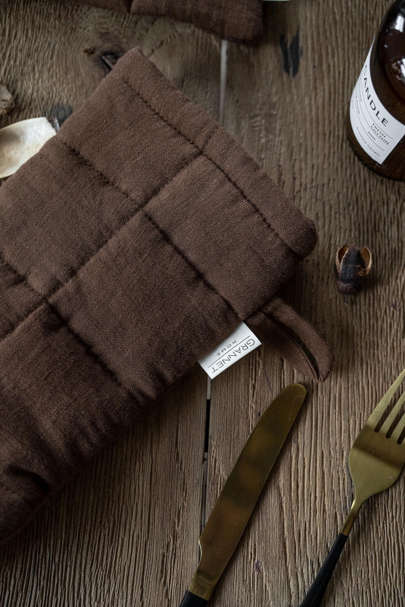 May include: A close-up of a brown quilted oven mitt with a loop for hanging. The mitt has a tag that reads "GRANNET HOME". A gold knife and fork are visible, along with a bottle and a small decorative item.