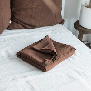 May include: A folded brown linen sheet on a white bed. The sheet is folded in thirds and has a smooth, natural texture.