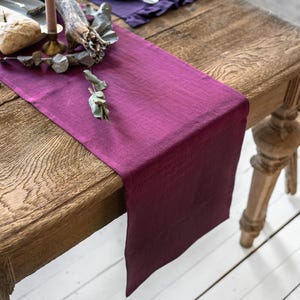 May include: A deep magenta rectangular table runner made of fabric with a visible weave texture. The runner is draped across a wooden table, extending over the edge. It features a solid, uniform color throughout its length and width.