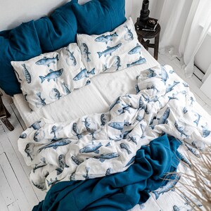 May include: A bed with white sheets, blue pillows, and a duvet cover featuring a blue fish pattern. A solid blue blanket is draped over the foot of the bed. The room has white walls and a wooden floor.
