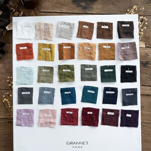 Pu&ograve; includere: Un pannello bianco con una collezione di campioni di tessuto di lino in vari colori. I campioni sono disposti in file e colonne, con etichette che indicano i nomi dei colori. Il pannello presenta il testo "GRANNET HOME" in basso.