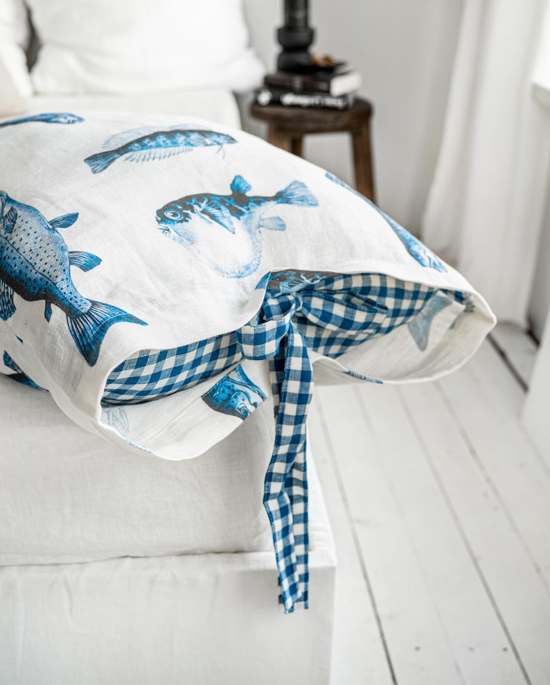 May include: White pillowcase with blue fish illustrations. The pillowcase has a blue and white gingham trim and a matching tie closure. The pillow is on a white bed.