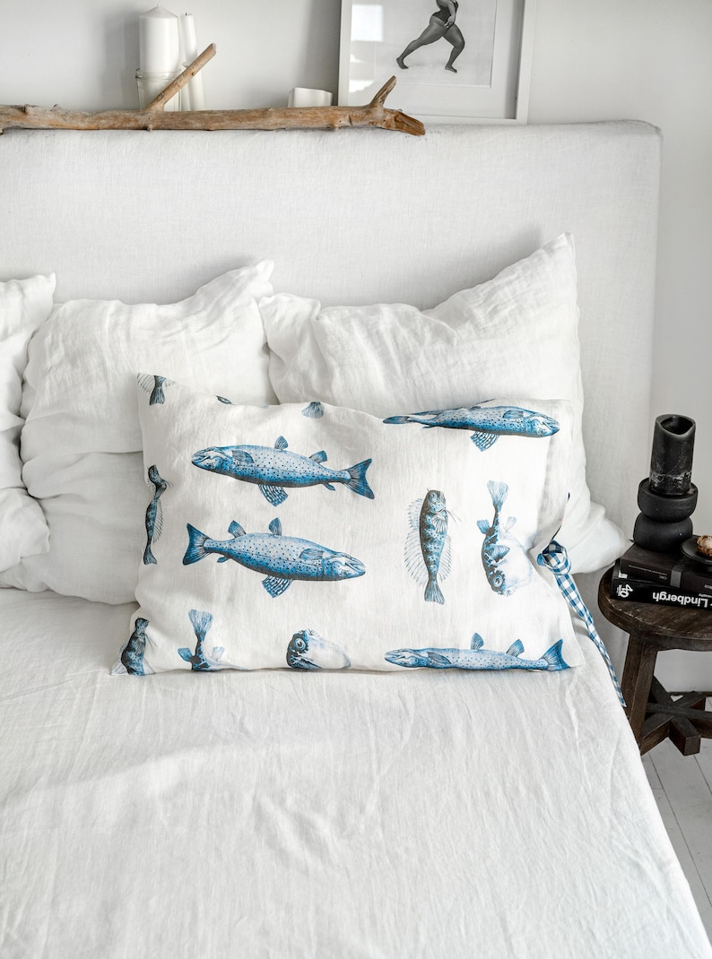 May include: A white rectangular pillow with a blue fish pattern rests on a white bed. The fish illustrations vary in size and orientation. The pillow is surrounded by white bedding and pillows, creating a serene and nautical theme.