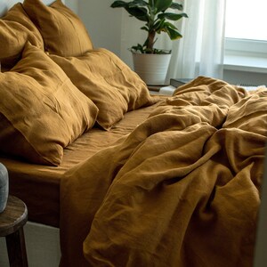 Puede incluir: Un juego de cama de lino amarillo mostaza con una funda n&oacute;rdica y varias fundas de almohada. La ropa de cama est&aacute; dispuesta sobre un marco de cama blanco con un taburete de madera en primer plano.