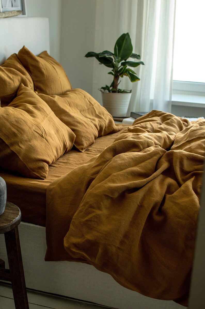Puede incluir: Un juego de cama de lino amarillo mostaza con una funda n&oacute;rdica y varias fundas de almohada. La ropa de cama est&aacute; dispuesta sobre un marco de cama blanco con un taburete de madera en primer plano.