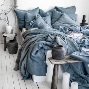May include: A bed with blue bedding, including pillows, a duvet, and a textured blanket. The bed is against a white headboard. Two small wooden tables hold decorative vases and candles. The floor is made of white painted wooden planks.