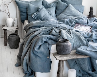 May include: A bed with blue bedding, including pillows, a duvet, and a textured blanket. The bed is against a white headboard. Two small wooden tables hold decorative vases and candles. The floor is made of white painted wooden planks.