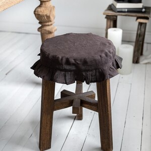 Pu&ograve; includere: Un piccolo sgabello in legno marrone scuro con una fodera rotonda in lino marrone scuro con volant. Lo sgabello ha quattro gambe robuste e una struttura di supporto a forma di croce. Sfondo con pavimento in legno verniciato di bianco.