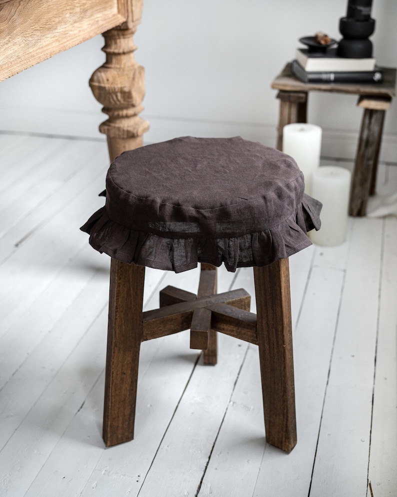 Pu&ograve; includere: Un piccolo sgabello in legno marrone scuro con una fodera rotonda in lino marrone scuro con volant. Lo sgabello ha quattro gambe robuste e una struttura di supporto a forma di croce. Sfondo con pavimento in legno verniciato di bianco.